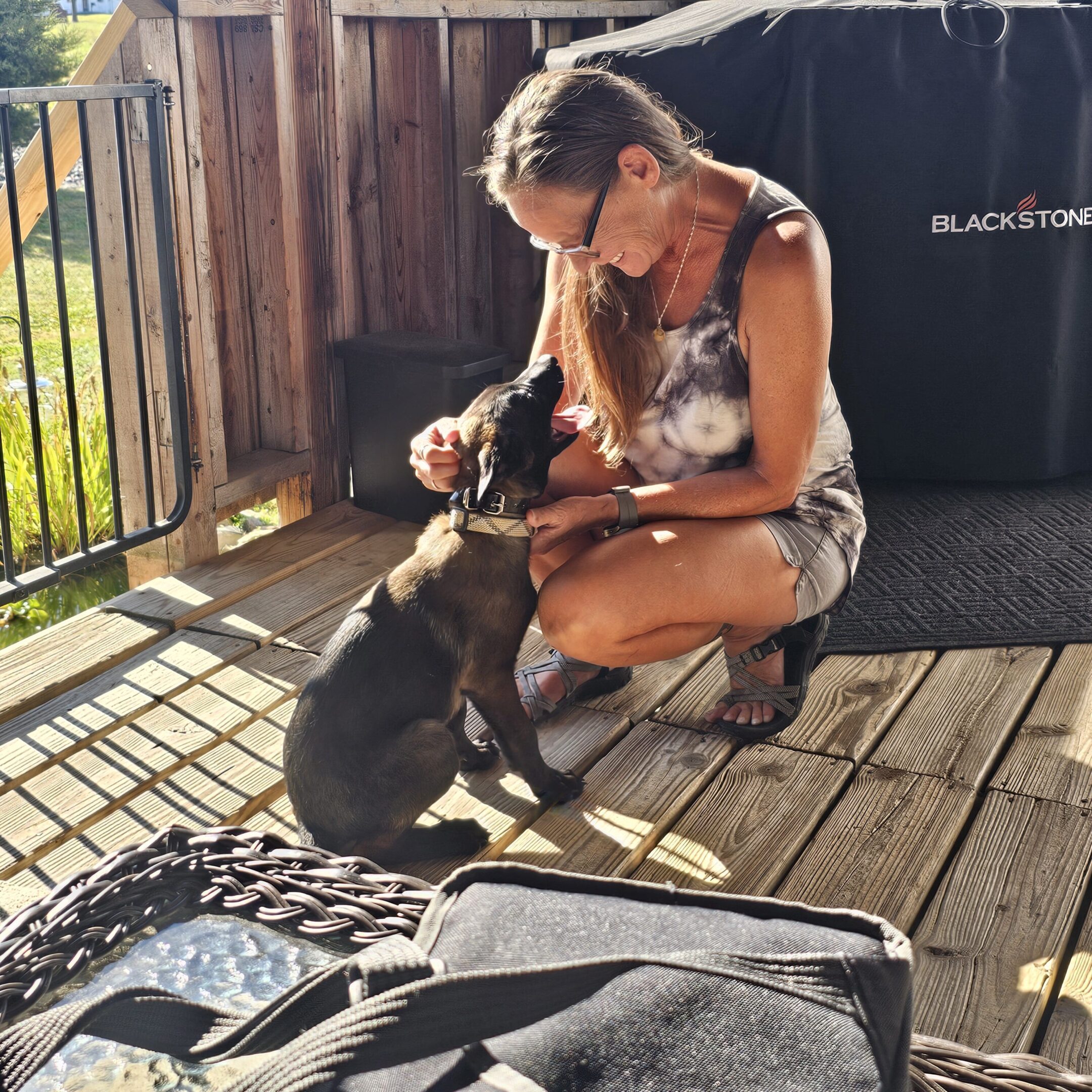 Woman affectionately petting a black dog on a wooden deck in sunlight.